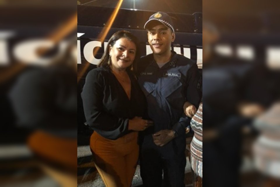 Mulher com blusa de manga comprida preta e calça marrom ao lado de homem com farda de policial