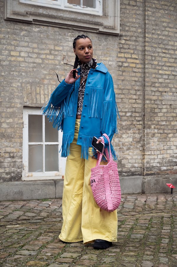 Mulher negra e jovem posando para foto nas ruas da cidade de Copenhagem, na Dinamarca. Ela tem os cabelos trançados e usa uma calça folgada amarela, uma blusa de oncinha, uma jaqueta azul de couro com franjas e uma bolsa de crochê rosa.