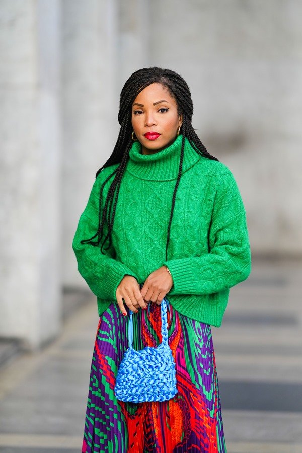 Mulher negra, jovem e com cabelos longos e trançados posando para foto na rua. Ela usa um casaco de crochê verde, uma saia estampada rosa, roxa e verde e uma bolsa de crochê azul.