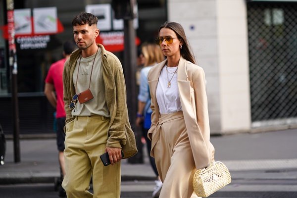 Mulher e homem, ambos jovens, brancos e com cabelos castanhos lisos, andando pela rua durante a Semana de Moda de Paris, na França. Ele usa uma camiseta cinza, calça bege e camisa de botão aberta por cima na cor verde militar. A mulher veste uma camiseta branca, um conjunto de blazer e calça bege, óculos escuros de lentes marrons e uma bolsa de palha.