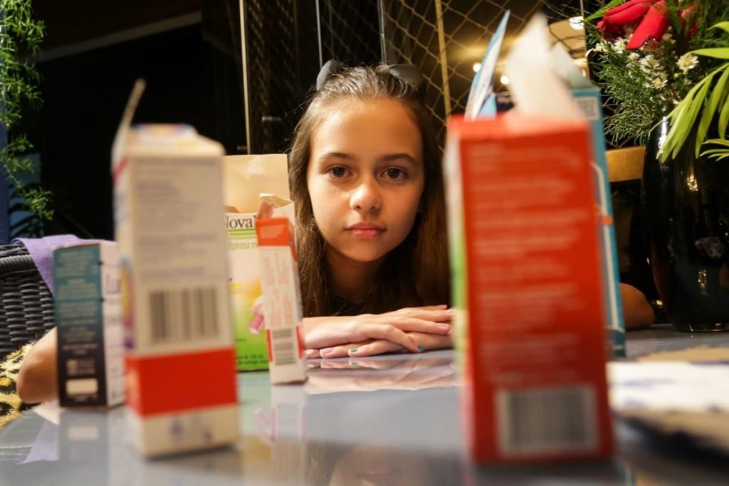 Menina branca de cabelos morenos olha entre caixas de remédios