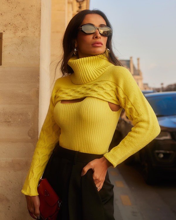 A influenciadora digital Silvia Braz, uma mulher branca com cabelo preto ondulado, posando para foto durante a Semana de Moda de Paris. Ela veste um tricô verde limão, calça preta, óculos escuros e bolsa caramelo