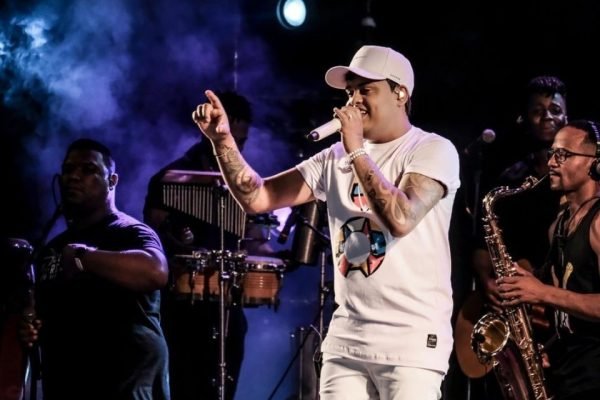 foto colorida mostra o cantor Thiago Aquino cantando em um palco com a sua banda