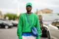 Homem negro e jovem posando para foto nas ruas de Paris, durante a semana de moda francesa. Ele usa uma camiseta roxa, suéter verde, bolsa transpassada roxa da Balenciaga, óculos prateado e um boné também verde.