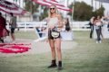 Mulher branca e jovem, com cabelos louros curtos e ondulados, posando para foto no festival de música Coachella, nos Estados Unidos. Ela está de óculos escuros e usa uma blusa bege com recorte ombro a ombro, um shorts jeans preto, um coturno de couro preto e uma bolsa transpassada branca da marca Chloé.