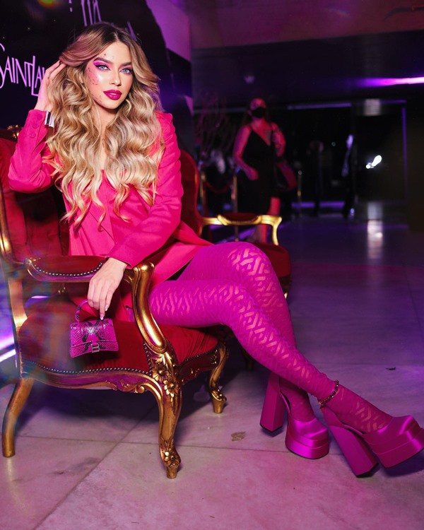 A influencer de moda Jordanna Maia, uma mulher jovem, branca, com cabelos louros, posando para foto em uma cadeira rosa. Ela usa um look em diferentes tons de rosa: meia calça, vestido, bolsa e sapato de salto alto.