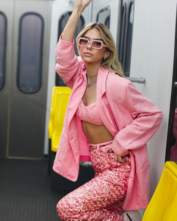 A influencer de moda Jordanna Maia, uma mulher jovem, branca, com cabelos louros, posando para foto em um vagão do metrô. Ela usa um look todo rosa claro: top curto, calça com lantejoulas e blazer. Usa, ainda, óculos escuros com armação brilhantes de estrasse.