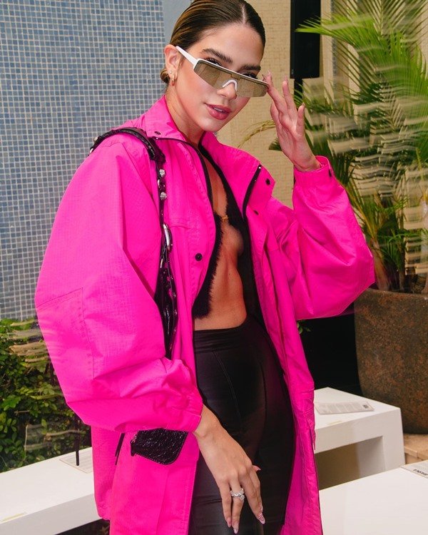 A influencer de moda Jordanna Maia, uma mulher jovem, branca, com cabelos louros, posando para foto na frente de um mosaico de cerâmica. Ela usa um top preto, um casaco rosa pink esportivo e óculos futurista prateado