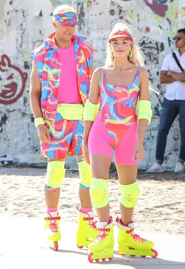 Actress Margot Robbie, a young, blond, white woman, shooting the movie Barbie with actor Ryan Gosling, a blond, white man.  They wear pink sportswear, with neon yellow accents, and skates at a skating rink.