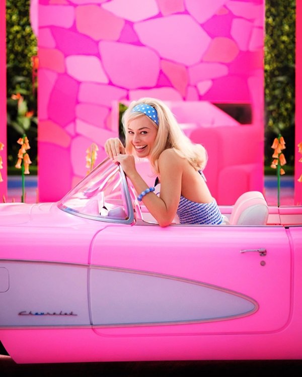 A atriz Margot Robbie, uma mulher branca, jovem e loura, gravando o filme Barbie. Ela está em um carro conversível rosa e usa um top e uma faixa na cabeça, ambos azuis com estampa de bolinha branca.