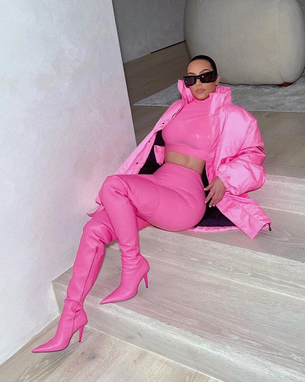 The businesswoman and influencer Kim Kardashian, a brunette woman with straight brown hair, posing for a photo on the marble stairs of her own residence.  She wears an all-pink outfit combination: crop top, pants, pointed toe boots, and padded sport coat.  She also wears black sunglasses.