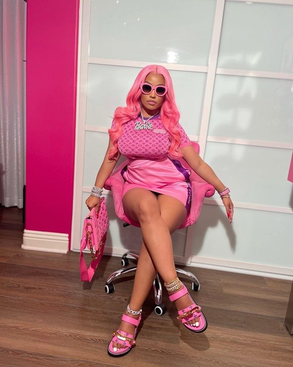 A cantora Nicki Minaj, uma mulher negra e jovem, sentada em uma cadeira rosa e usando peruca da mesma cor. Ela usa toda uma combinação de roupas em diferentes tons da cor rosa: vestido, sandália de salto, bolsa e óculos