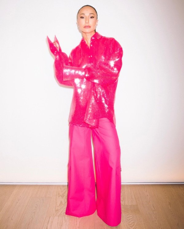 Presenter and former BBB Sabrina Sato, a young, brunette woman with brown hair and Asian physical features, posing for a photo on a white wall.  She wears a pink sequined shirt, baggy pink pants, and pink gloves in the same color.