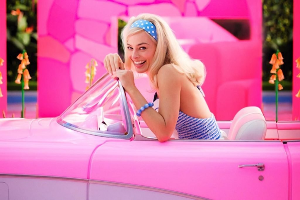Actress Margot Robbie, a young, blond, white woman, filming the movie Barbie.  She is in a pink convertible car and wears a top and headband, both blue with a white polka dot print.