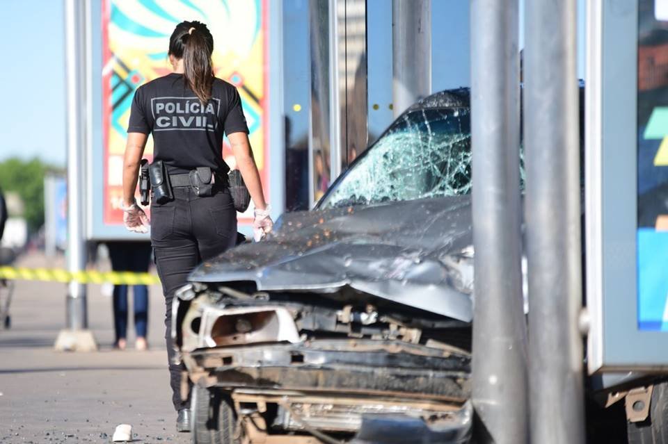 Carro cinza amassado encostado em poste e mulher com roupa preta de costas ao lado