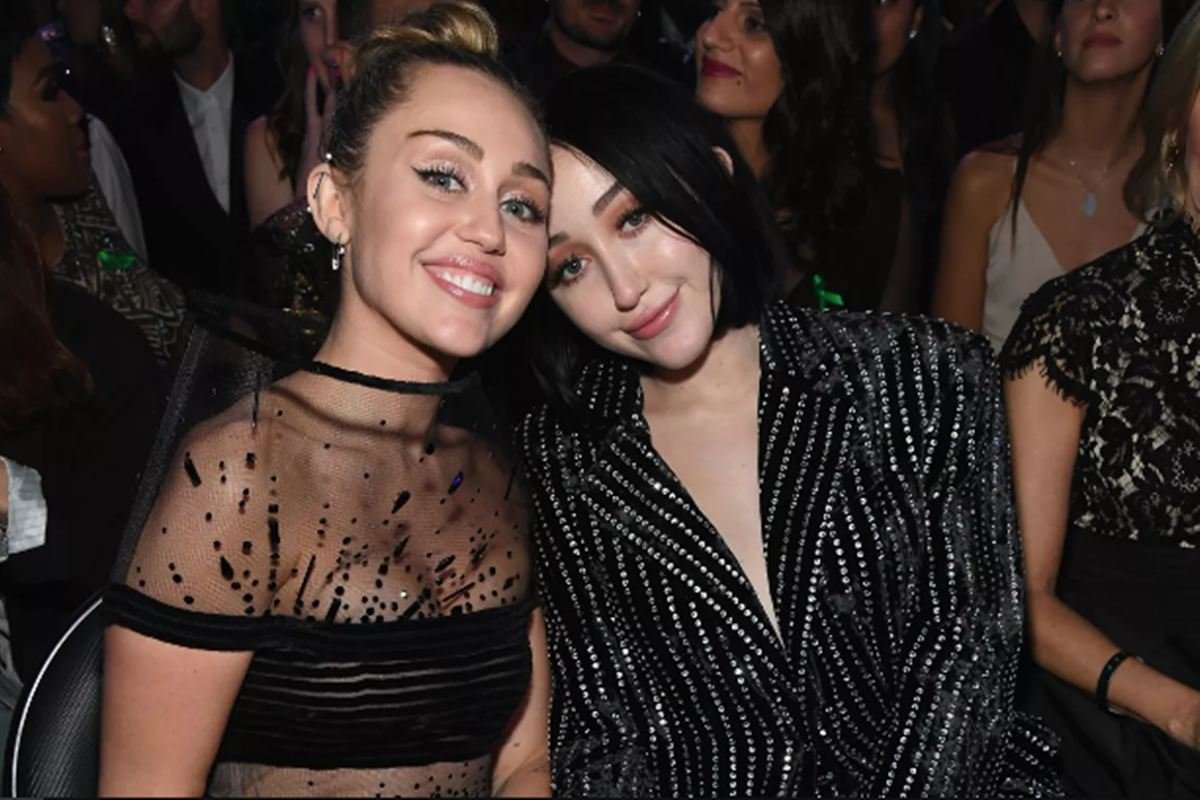 Miley Cyrus e irmã, Noah Cyrus, em premiação. Ambas sentam-se na plateia e sorriem com os rostos encostados. Elas usam roupas pretas com brilho - Metrópoles