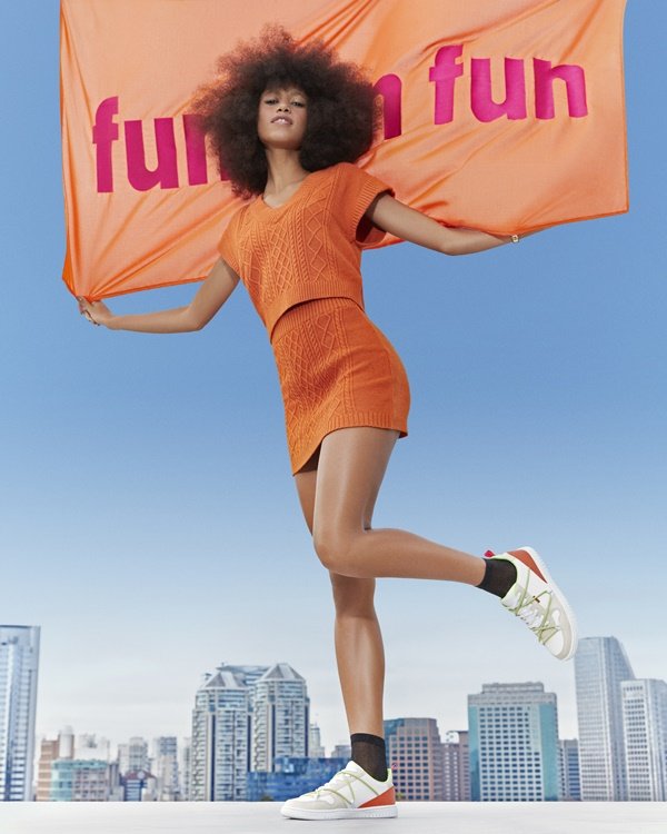 Campanha de divulgação da nova coleção da marca Anacapri. A modelo que posa para foto, uma mulher jovem, negra e com cabelo black power, usa conjunto de tricô laranja e tênis e bolsa da marca.