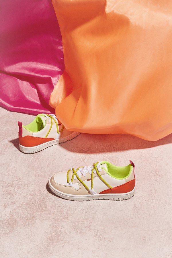 Campanha de divulgação da nova coleção da marca Anacapri. Na foto é possível ver um tênis branco da marca, com detalhes vermelhos e verde limão, em cima de tecidos laranjas e rosas.
