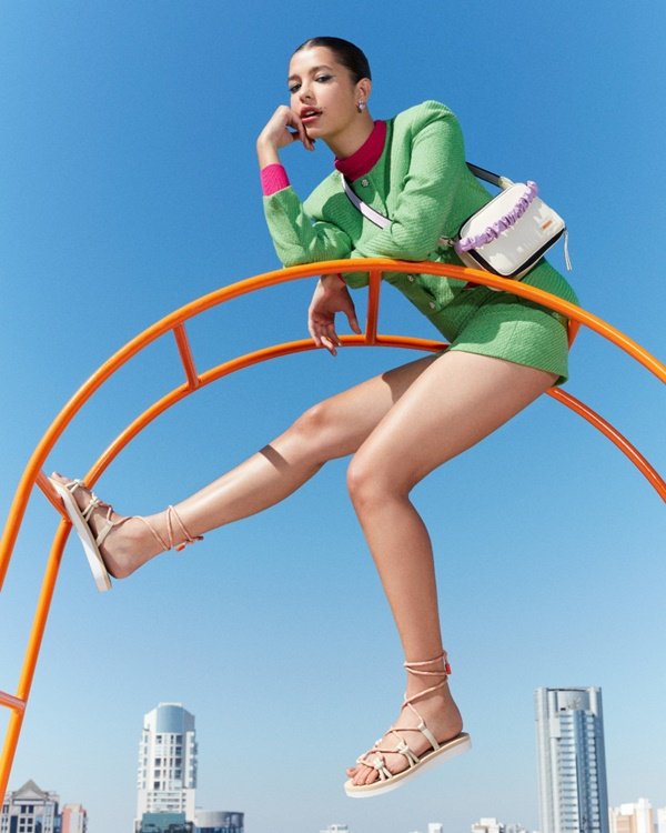 Campanha de divulgação da nova coleção da marca Anacapri. A modelo que posa para foto, uma mulher jovem, branca e com cabelo liso castanho, com um conjunto de shorts e blusa de manga verde, uma bolsa pequena branca e sandálias estilo gladiadora branca.