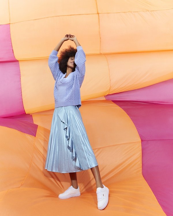 Campanha de divulgação da nova coleção da marca Anacapri. A modelo que posa para foto, uma mulher jovem, negra e com cabelo black power, usa saia plissada azul brilhante, tricô lilás de crochê e tênis branco da marca.