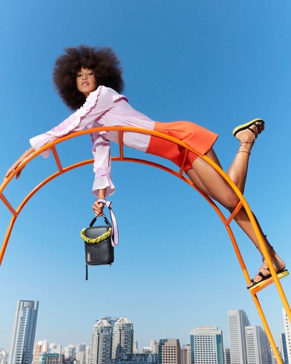 Campanha de divulgação da nova coleção da marca Anacapri. A modelo que posa para foto, uma mulher jovem, negra e com cabelo black power, usa blusa de manga cumprida branca, saia laranja, sandália rasteira e uma bolsa pequena preta.