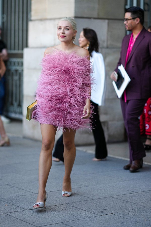 Mulher brancae jovem, com cabelo curto e louro penteado para trás, na Semana de Alta-Costura em Paris. Ela usa um vestido rosa e curto de plumas, uma bolsa dourada na mão e uma sandália de salto prateada