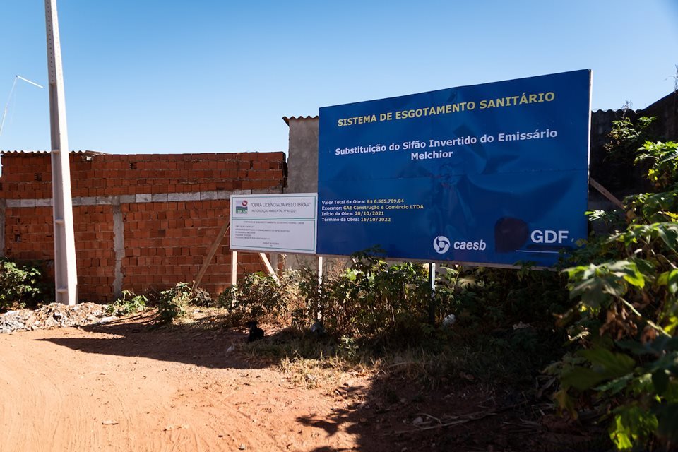 Placa do Governo do Distrito Federal anuncia sistema de esgotamento sanitário próxima a casas de alvenaria pequenas construídas em descampado numa invasão do Sol Nascente próxima à parque - Metrópoles