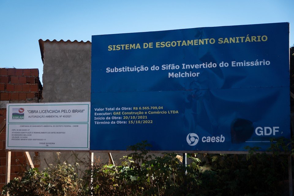 Placa do Governo do Distrito Federal anuncia sistema de esgotamento sanitário próxima a casas de alvenaria pequenas construídas em descampado numa invasão do Sol Nascente próxima à parque - Metrópoles