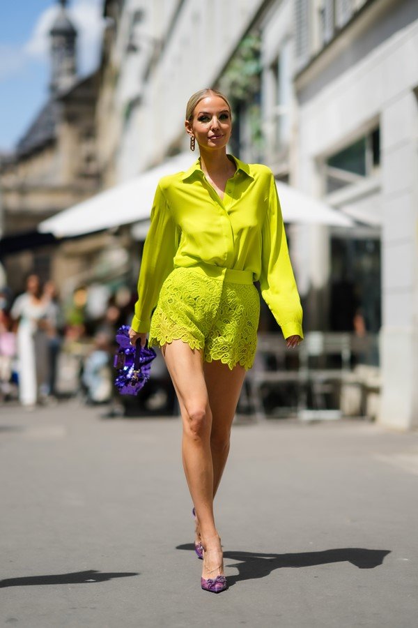 Mulher branca e jovem, com cabelo louro amarrado em um coque, andando pelas ruas de Paris durante a Semana de Alta-Costura. Ela usa um conjunto de camisa de botão e shorts de renda, tudo em um tom de verde limão neon, e sandália roxa de salto alto.