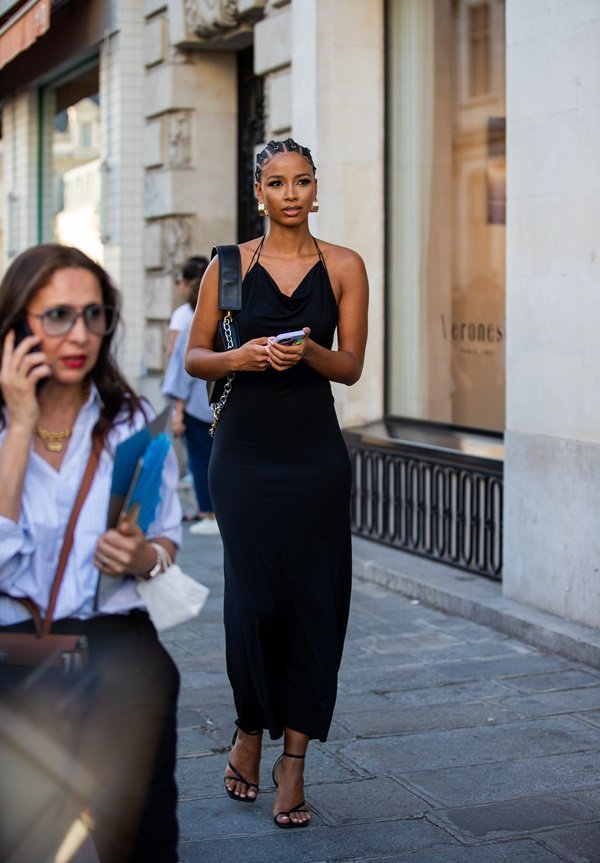 Mulher negra e jovem, com cabelo trançado, andando pelas ruas de Paris durante a Semana de Alta-Costura. Ela usa um vestido de cetim justo e longo, que lembra uma camisola, com alcinhas, e uma sandália de salto de tiras pretas.
