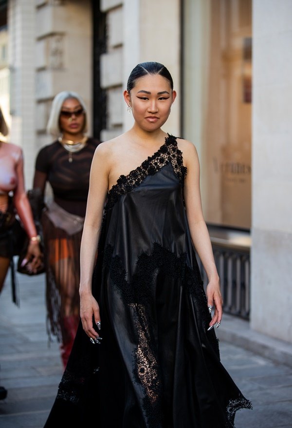 Mulher jovem e asiática, com cabelo preto amarrado, andando pelas ruas de Paris durante a Semana de Alta-Costura. Ela usa um vestido preto com detalhes de renda e bordados. O materiald a peça é brilhante e lembra uma lona.
