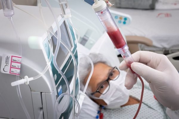 Mulher com cabelos brancos e óculos deitada em cama de hospital olhando para tubo com sangue ao lado de equipamento