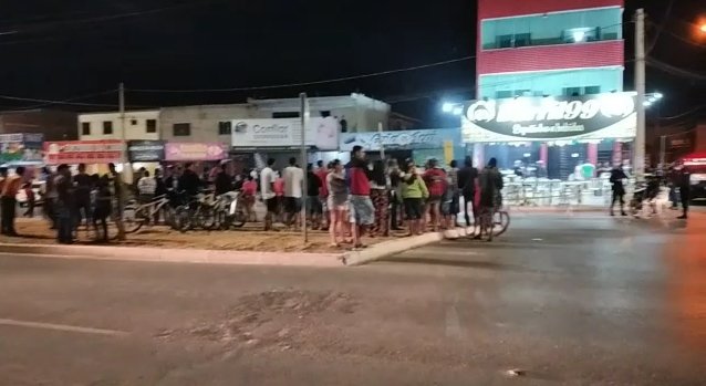 Foto colorida. Multidão observa corpo de homem morto a tiros
