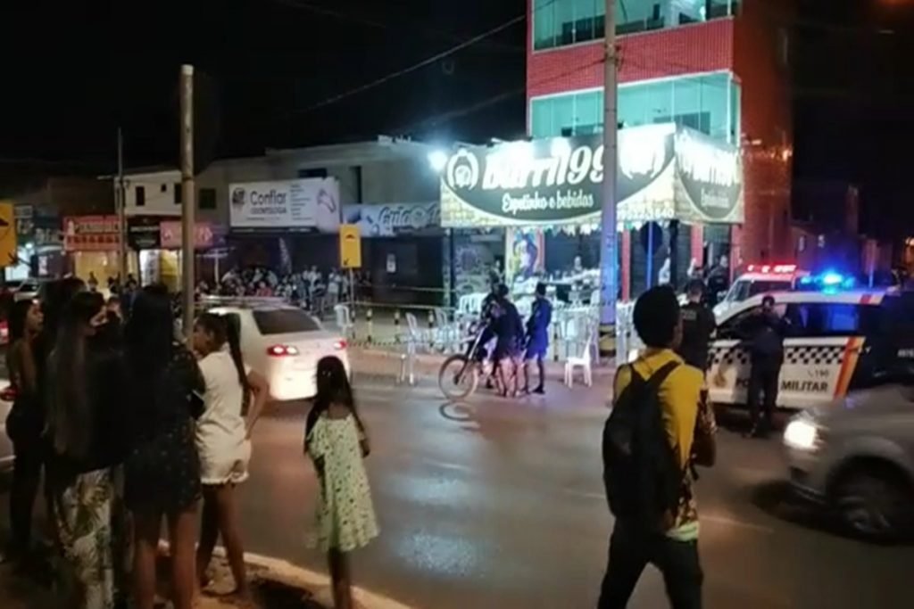 Foto colorida. Multidão observa corpo de homem morto a tiros