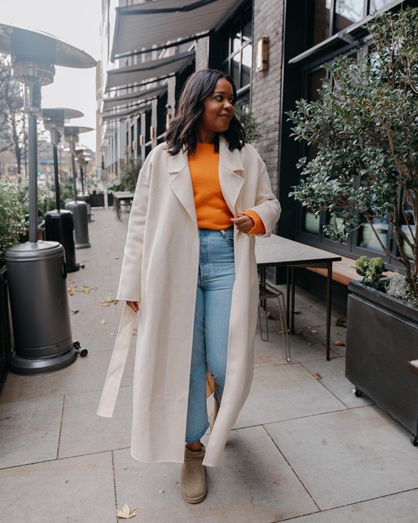 Mulher jovem e negra, de cabelo médio ondulado, andando pelas ruas de Londres. Ela usa uma camiseta laranja, calça jeans clara, uma bota marrom e um trech coat bege.