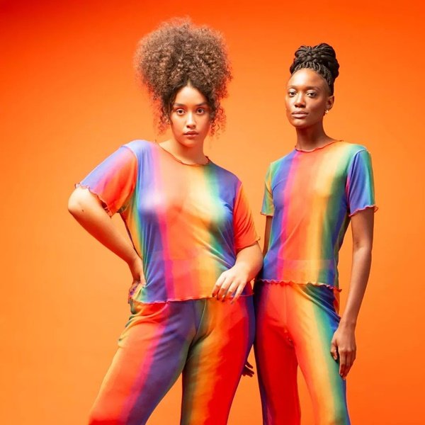 Duas modelos negras vestindo a mesma roupa da C&A: um conjunto de blusa e calça com listras que lembram um arco-iris. A primeira é uma menina de manequim tamanho médio e a segunda é magra.