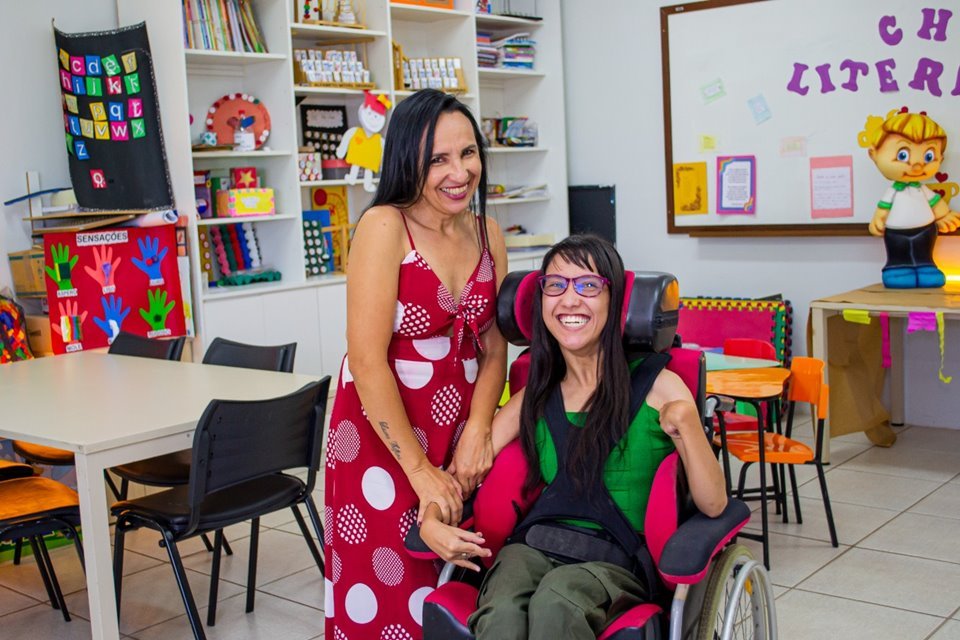 Mulher com vestido rosa em pé ao lado de menina em cadeira de rodas. Elas estão dentro de uma sala de aula colorida