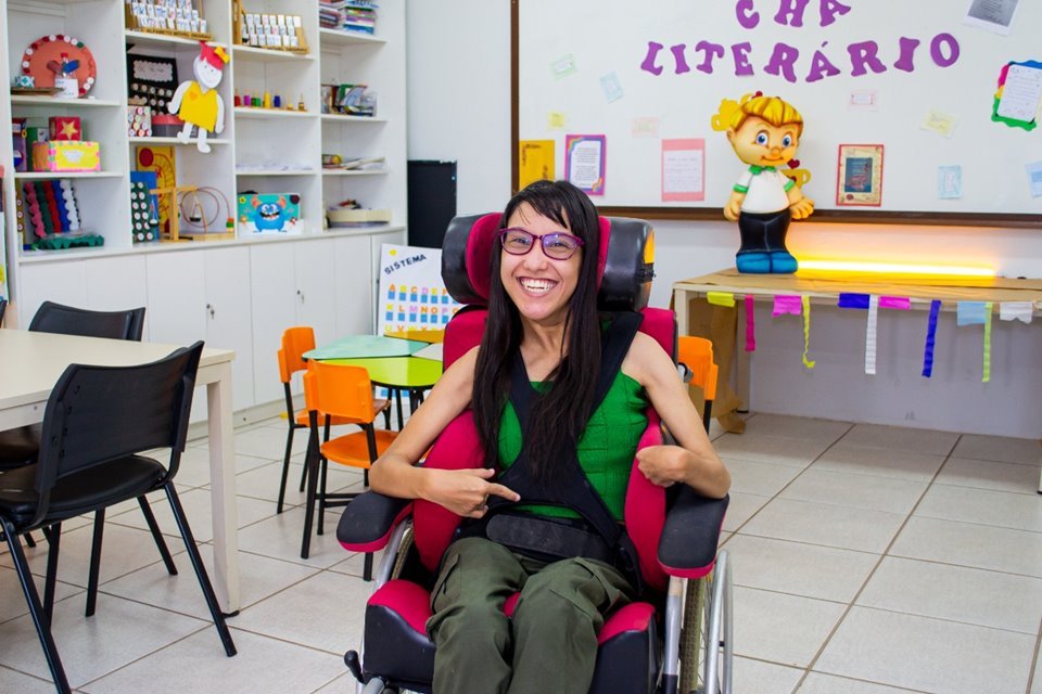 Menina com camisa verde e calça cinza sentada em cadeira de rodas. Ela está em uma sala de aula colorida