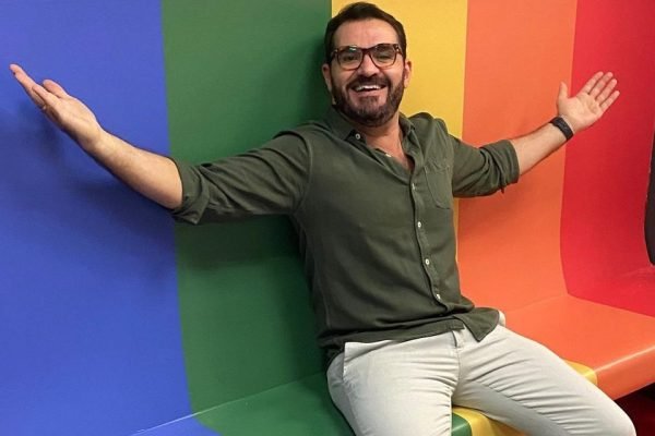 O repórter Marcelo Cosme, da TV Globo, posa sorrindo e de braços abertos diante de uma parede de arco-íris. Ele denunciou comentário homofóbico no seu Instagram - Metrópoles