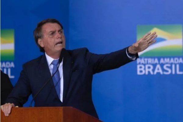 Bolsonaro de pé e vestindo terno e gravata. Ele fala em um microfone e aponta para o lado - Metrópoles