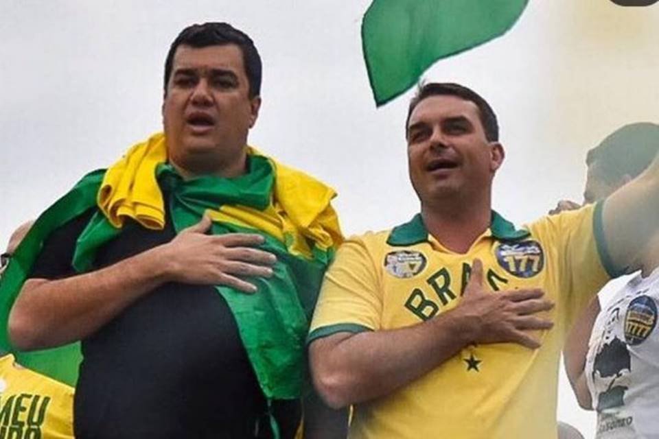 O senador Flávio Bolsonaro (direita) e seu suplente, Léo Rodrigues. Ambos aparecem em carro de som, numa manifestação governista, cantando o hino nacional com a mão no peito. Eles usam as cores da bandeira do Brasil - Metrópoles