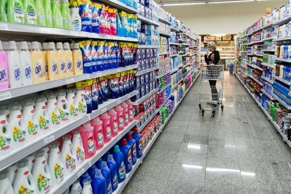 Mulher branca e loira olha prateleiras de produtos de limpeza no supermercado enquanto segura carrinho com dois detergentes dentro - Metrópoles, alimentação