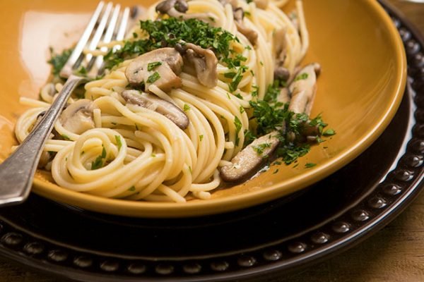 Espaguete com cogumelos salpicado com ervas em prato amarelo com garfo - Metrópoles
