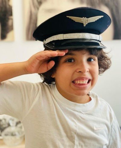 Menino com camisa branca sorrindo e com a mão na cabeça. Ele usa um chapéu azul de piloto