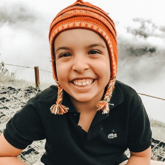 Menino com camisa preta e touca laranja sorrindo com nuvens atrás