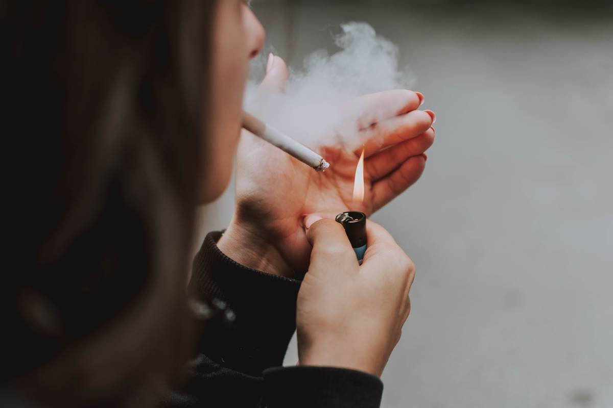 Mulher acendendo cigarro e fumando, segurando um isqueiro e contendo a fumaça com a outra mão. Ela é visto de costas - Metrópoles