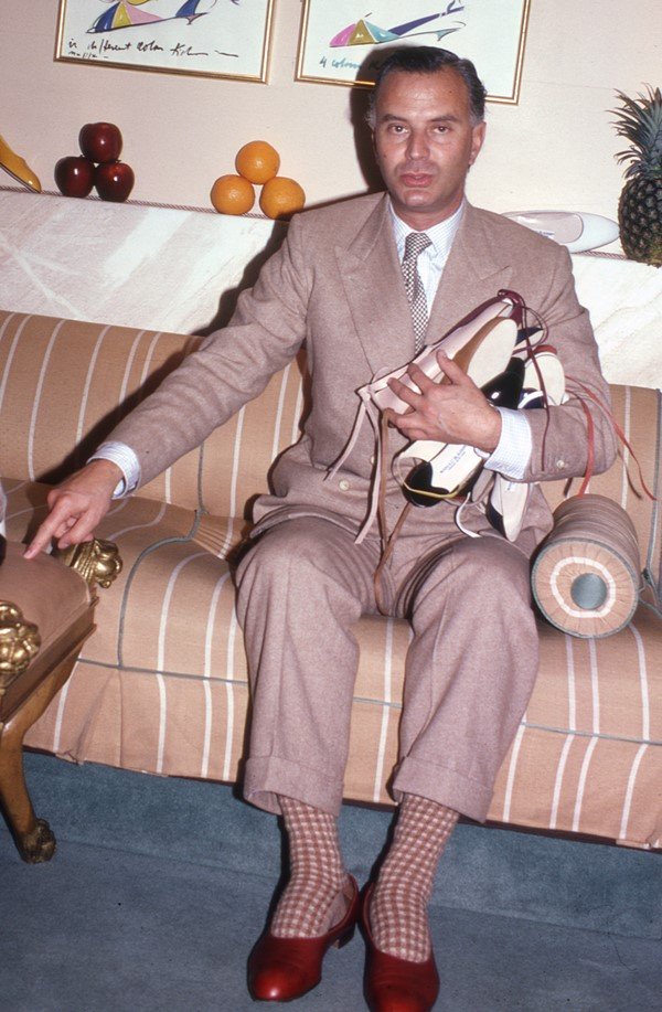 O estilista Manolo Blahnik, um homem branco e de meia idade, sentado em um sofá bege segurando sandálias que ele mesmo desenvolveu. Veste um terno bege e usa uma camisa branca com gravata cinza