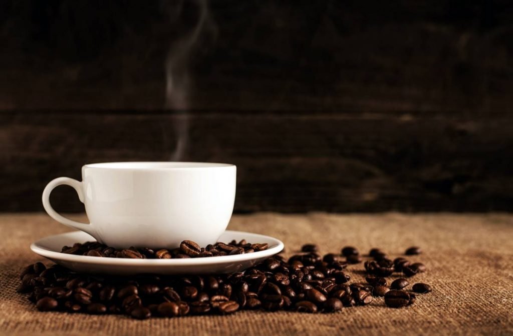 Xícara de café com fumaça saindo sob tecido marrom e vários grãos sob pires - Metrópoles