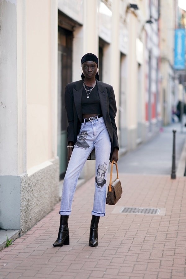 Mulher jovem e negra posando para foto nas ruas de Paris durante a semana de moda francesa. Ela usa um lenço preto amarrado na cabeça, blusa e blazer também pretos, calça jeans clara e bota de couro.
