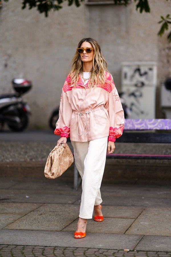 Mulher branca e jovem, com cabelos loiros e ondulados, andando nas ruas de Paris durante a semana de moda francesa. Ela usa camiseta e calça, ambas peças brancas, um casaco estilo corta-vendo rosa e uma bolsa também rosa.